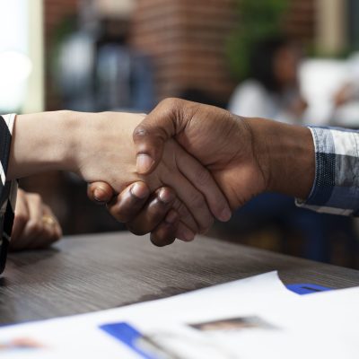 Closeup of multiethnic individuals handshaking over the office desk during job interview meeting. Selective focus on hands of diverse people doing a greeting gesture in brick wall workspace.